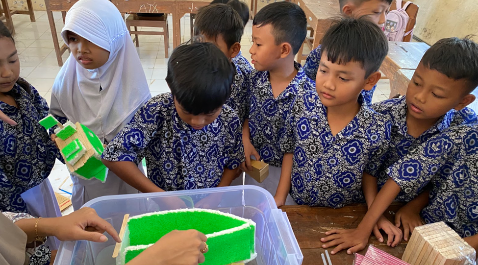 Sekelompok siswa SD berseragam batik biru sedang fokus mengamati eksperimen benda terapung berwarna hijau di dalam wadah plastik berisi air.