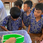 A group of elementary school students in blue batik uniforms focused on observing a green floating object experiment in a plastic water tub.