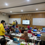 A training atmosphere inside a modern classroom where dozens of participants are sitting at their desks, using laptops, and looking at a presentation screen at the front.