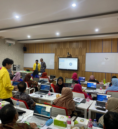 A training atmosphere inside a modern classroom where dozens of participants are sitting at their desks, using laptops, and looking at a presentation screen at the front.