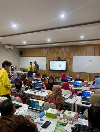 A training atmosphere inside a modern classroom where dozens of participants are sitting at their desks, using laptops, and looking at a presentation screen at the front.