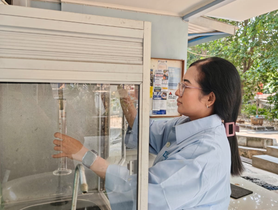 Seorang perempuan berkacamata dan mengenakan kemeja biru muda sedang memegang tabung ukur kaca di dekat keran air dan wastafel.