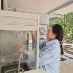 A woman with glasses wearing a light blue shirt holding a glass measuring cylinder near a water tap and sink.