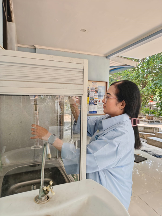 A woman with glasses wearing a light blue shirt holding a glass measuring cylinder near a water tap and sink.