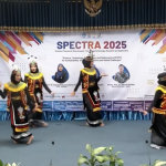 Lima mahasiswi mengenakan kostum tari tradisional berwarna hitam dengan aksen emas dan hiasan kepala sedang menari di atas panggung berlatar belakang spanduk acara SPECTRA 2025.