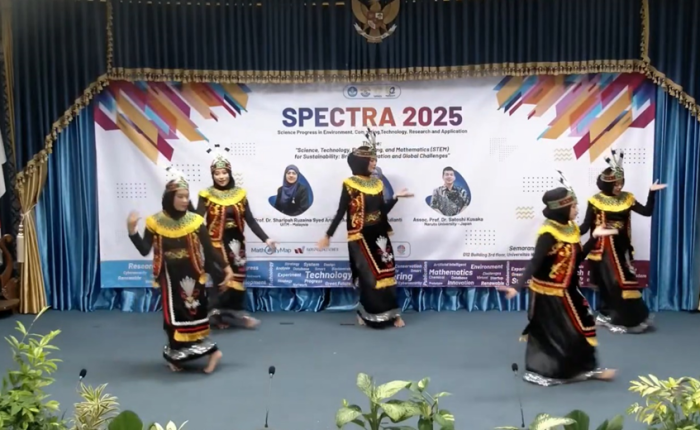 Five female students wearing traditional black dance costumes with gold accents and headpieces performing on a stage with a SPECTRA 2025 event banner in the background.