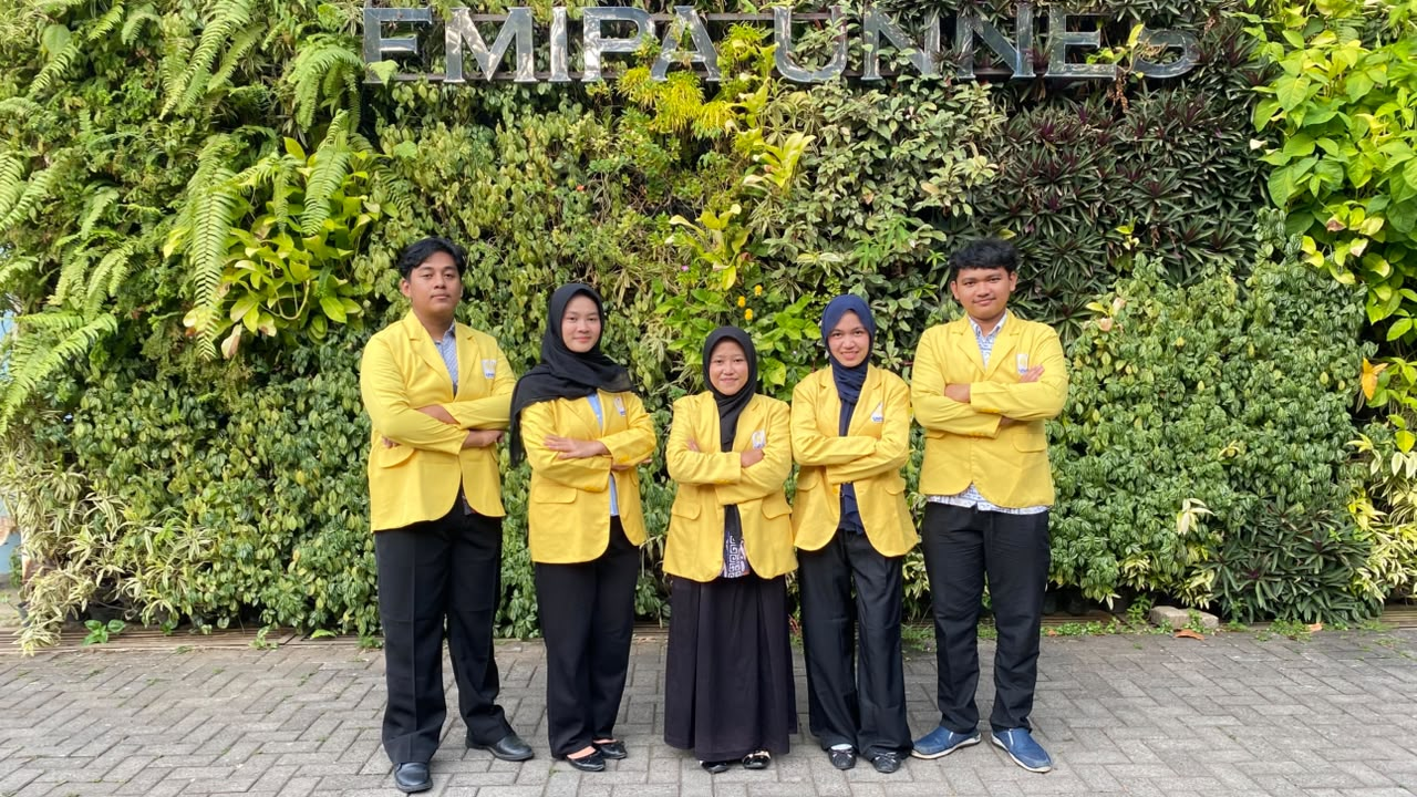 Lima mahasiswa yang terdiri dari dua laki-laki dan tiga perempuan mengenakan jas almamater kuning kuning, berpose melipat tangan di dada dengan latar belakang taman vertikal hijau bertuliskan FMIPA UNNES.