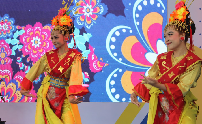 Dua penari perempuan mengenakan kostum tradisional berwarna kuning dan merah dengan hiasan kepala emas bermotif bunga, menari di atas panggung dengan latar belakang layar digital bermotif bunga warna-warni.