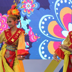 Two female dancers wearing yellow and red traditional costumes with golden floral headdresses, performing on a stage with a colorful floral patterned digital screen in the background.