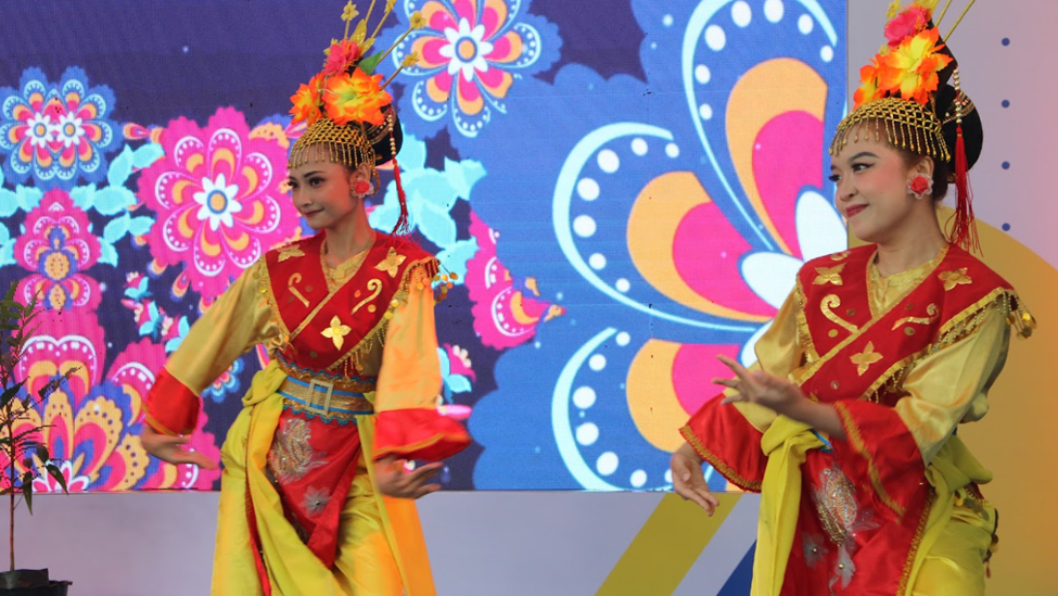 Two female dancers wearing yellow and red traditional costumes with golden floral headdresses, performing on a stage with a colorful floral patterned digital screen in the background.