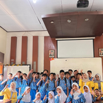 Sekelompok mahasiswa perempuan mengenakan jas almamater kuning khas UNNES berpose ceria bersama puluhan murid SMP berseragam biru putih di dalam ruang kelas.