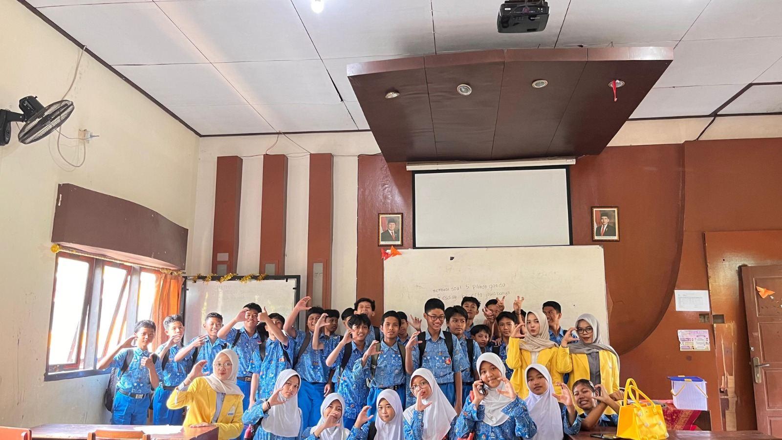 Sekelompok mahasiswa perempuan mengenakan jas almamater kuning khas UNNES berpose ceria bersama puluhan murid SMP berseragam biru putih di dalam ruang kelas.