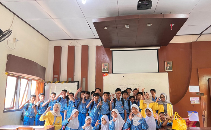 A group of female university students wearing UNNES signature yellow alma mater jackets posing cheerfully with dozens of junior high school students in blue and white uniforms inside a classroom.