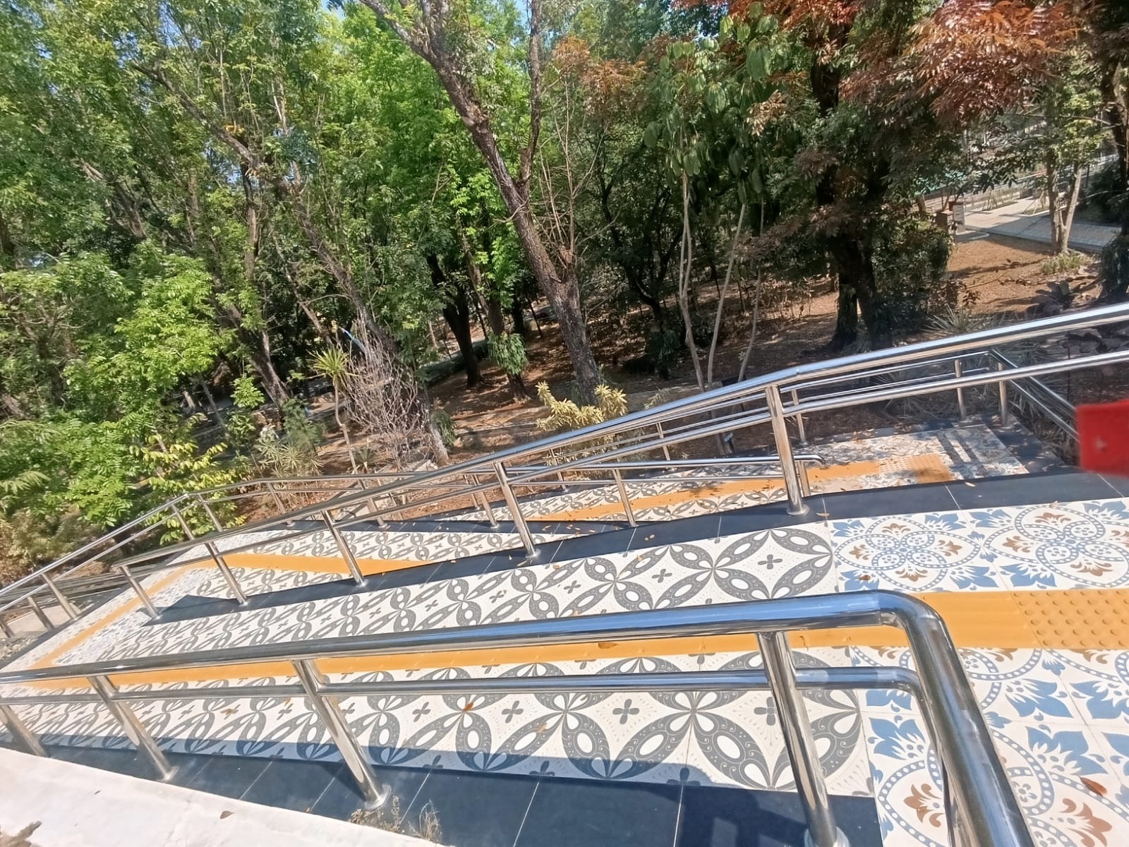 A patterned tiled ramp with yellow tactile paving in the center and stainless steel handrails on both sides in the outdoor area of the FMIPA UNNES building surrounded by green trees.