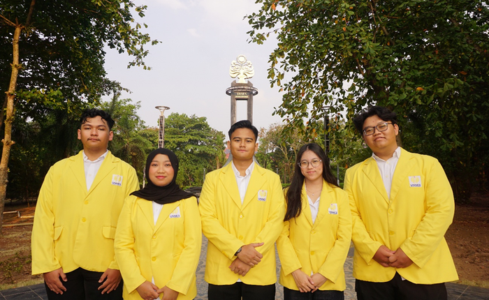 Five students consisting of three males and two females wearing the signature UNNES yellow alma mater jackets, standing in a row facing the camera outdoors with trees and the campus conservation monument in the background.