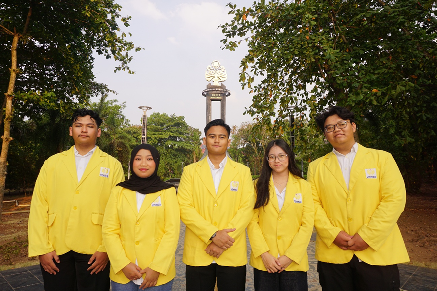Five students consisting of three males and two females wearing the signature UNNES yellow alma mater jackets, standing in a row facing the camera outdoors with trees and the campus conservation monument in the background.