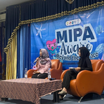 Dua orang perempuan berhijab sedang duduk di atas panggung menggunakan kursi sofa berwarna oranye. Salah satu perempuan sedang berbicara menggunakan mikrofon, sementara satu lainnya menyimak dengan membawa catatan. Di belakang mereka terdapat spanduk besar berwarna biru bertema bawah laut dengan tulisan MIPA Aware Beyond the Surface.