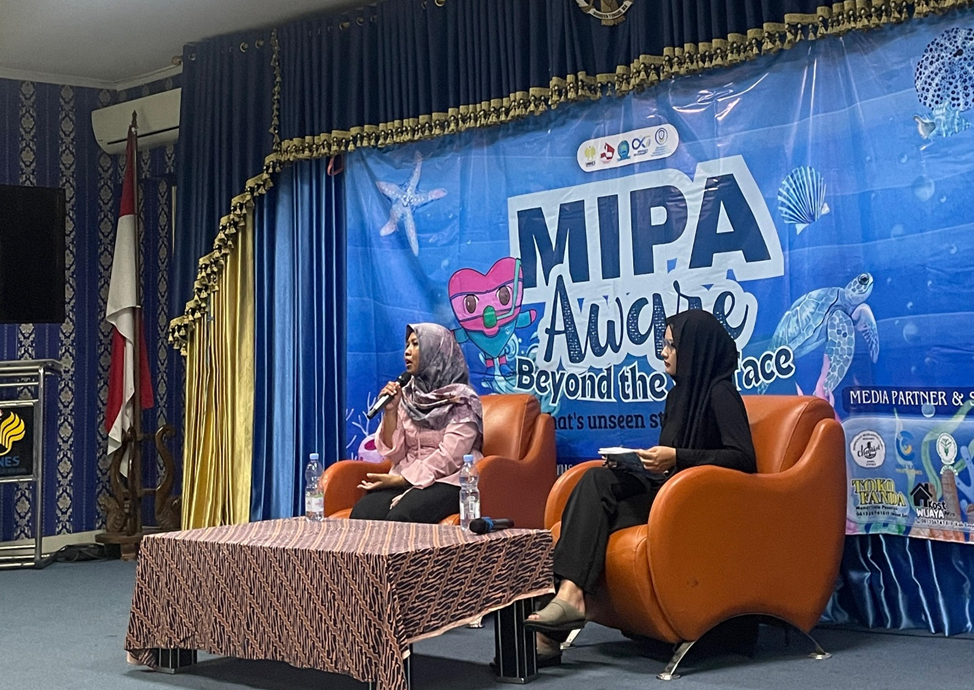 Dua orang perempuan berhijab sedang duduk di atas panggung menggunakan kursi sofa berwarna oranye. Salah satu perempuan sedang berbicara menggunakan mikrofon, sementara satu lainnya menyimak dengan membawa catatan. Di belakang mereka terdapat spanduk besar berwarna biru bertema bawah laut dengan tulisan MIPA Aware Beyond the Surface.