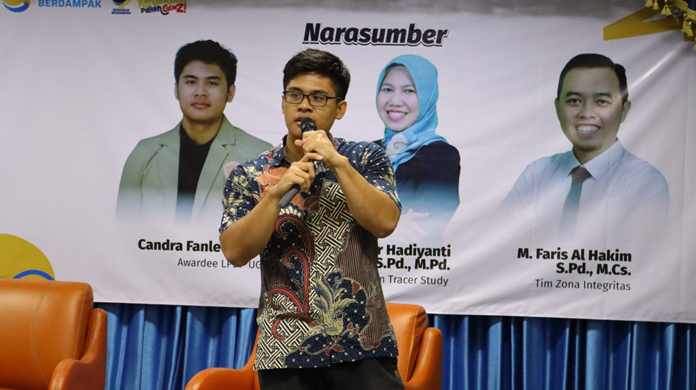 Narasumber Candra Fanleon berdiri memegang mikrofon di depan panggung dengan latar belakang spanduk yang menampilkan foto tiga narasumber workshop karier FMIPA UNNES.