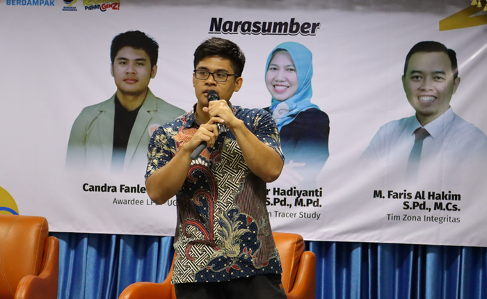 Speaker Candra Fanleon standing while holding a microphone in front of a stage with a background banner featuring photos of the three career workshop speakers at FMIPA UNNES.