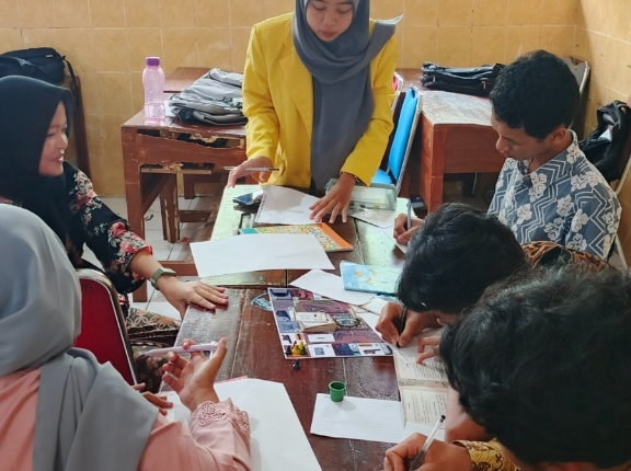 Seorang mahasiswa perempuan mengenakan jas almamater kuning UNNES mendampingi empat murid SMP yang sedang duduk melingkar mengerjakan tugas di meja kayu di dalam kelas.
